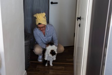 Koridorun ortasında elinde beyaz ve kahverengi bir kedi tutan biri oturuyor. Kafalarında bir havlu var ve günlük kıyafetler giyiyorlar. Gündüz ve içerisi aydınlık.