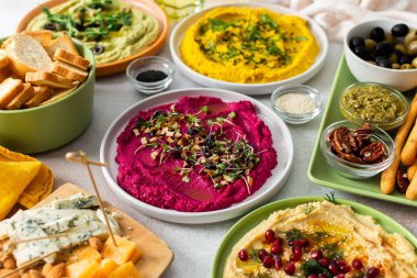Renkli bir şarküteri tahtasında pancar kökü humus, klasik humus, brokoli humusu ve sosla süslenmiş, ekmek ve zeytinle çevrili zerdeçal humus yer alıyor..