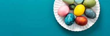Paskalya yumurtalarının çeşitli renkleri ve desenleri parlak mavi arka planda yuvarlak beyaz bir tabakta sergilenir. Bayram kutlamaları ve afişler için mükemmeldir.