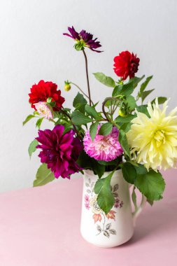 Bir vazo farklı renklerde taze çiçekler içerir. Çiçekler pembe bir masanın üzerine konur. Vazonun arkasında parlak bir odada düz bir duvar var.