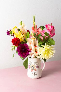 Bir vazo farklı renklerde taze çiçekler içerir. Çiçekler pembe bir masanın üzerine konur. Vazonun arkasında parlak bir odada düz bir duvar var.