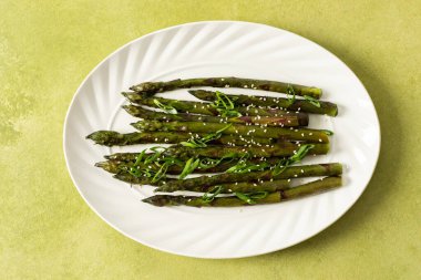 Taze ızgara kuşkonmaz, üzerinde susam tohumları ve taze otlar bulunan beyaz bir tabakta, rahat bir mutfakta yeşil bir arka planda bekletiliyor.