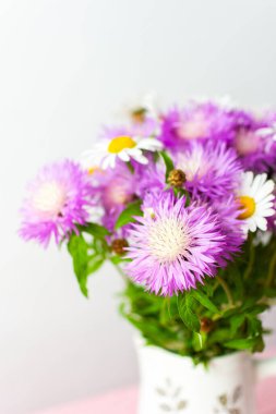 Renkli bir buket papatya ve mor çiçekleri büyüleyici bir sürahiye yerleştirilmiş pembe bir masa örtüsünün üzerinde duruyor. Yumuşak ve sade ortama neşe katıyor.