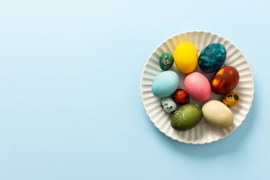 Süslü Paskalya yumurtalarının çeşitli renkleri, bahar şenlikleri ve neşesini sembolize eden yumuşak mavi arka planda beyaz bir tabağa yerleştirilmiştir.