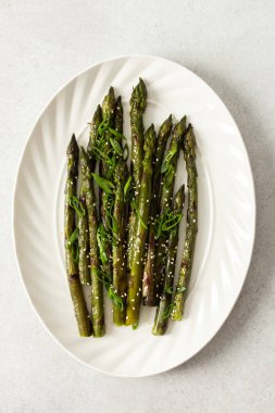 Taze otlarla kaplı buharda pişirilmiş kuşkonmaz, beyaz bir tabakta güzelce sıralanır. Yemek canlı görünüyor ve sağlıklı bir yemek ya da yan yemek için uygun.