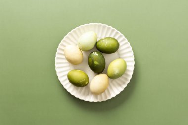 Yeşil ve soluk renklerin çeşitli tonlarında güzel boyanmış Paskalya yumurtaları, bahar mevsimi boyunca neşe ve yenilenmeyi sembolize eden beyaz bir tabakta duruyor.