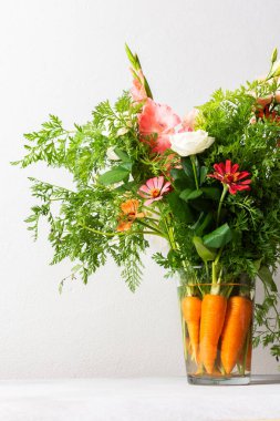 Bir buket gül, gladioli, zinya ve Eustomas suyun içinde duruyor. Yeşil uçlu havuçlar, açık arkaplana karşı düz bir masa üzerindeki düzenlemenin bir parçasıdır.