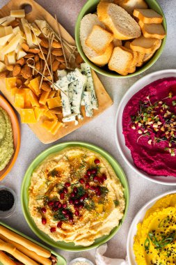 Renkli bir şarküteri tahtasında pancar kökü humus, klasik humus, brokoli humusu ve sosla süslenmiş, ekmek ve zeytinle çevrili zerdeçal humus yer alıyor.