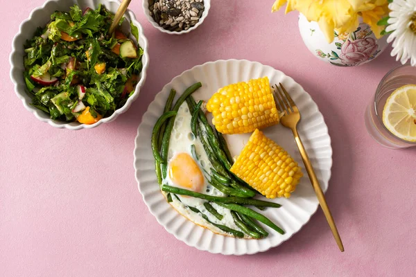 Sağlıklı bir kahvaltı pembe bir masada servis edilir. İçinde kuşkonmaz, sahanda yumurta, mısır ve taze salata var. Üzüm ve ayçekirdeği yan tarafta.