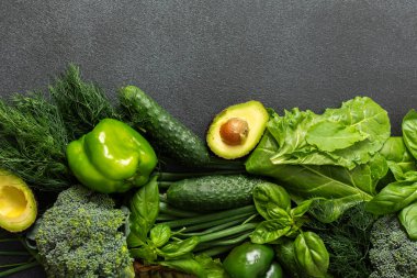 A vibrant assortment of fresh green vegetables is carefully arranged on a dark surface. This selection includes broccoli, green bell peppers, cucumbers, spinach, lettuce, and an avocado, healthy eating