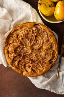 Taze bir armut turtası masanın üzerindeki bir beze yerleştirilir. Yakınlarda olgun armutlar bulunan bir kase ve küçük bir vazonun çiçekleri var. Ortam sıcak ve ikram için davetkar görünüyor.