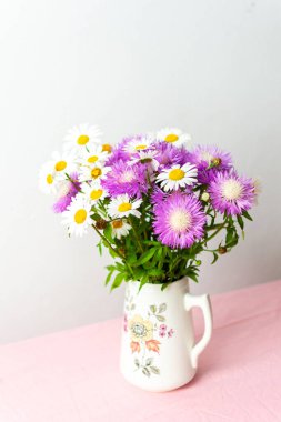 Renkli bir buket papatya ve mor çiçekleri büyüleyici bir sürahiye yerleştirilmiş pembe bir masa örtüsünün üzerinde duruyor. Yumuşak ve sade ortama neşe katıyor.