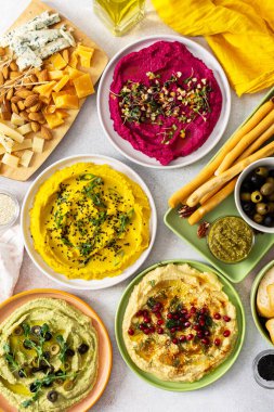 A variety of hummus flavors including turmeric, classic, and broccoli are served with fresh bread, nuts, and garnishes on a colorful charcuterie board, perfect for sharing