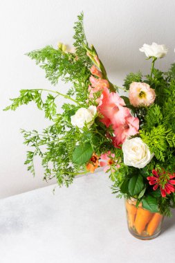 Bir buket gül, gladioli, zinya ve Eustomas suyun içinde duruyor. Yeşil uçlu havuçlar, açık arkaplana karşı düz bir masa üzerindeki düzenlemenin bir parçasıdır.