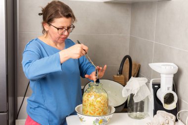 Bir kadın mutfakta duruyor, mayalanmak için lahana karıştırıyor. Kavanozları depoya hazırlarken lahana karıştırmak için kaşık kullanıyor. Atmosfer yemek hazırlıklarıyla dolu.