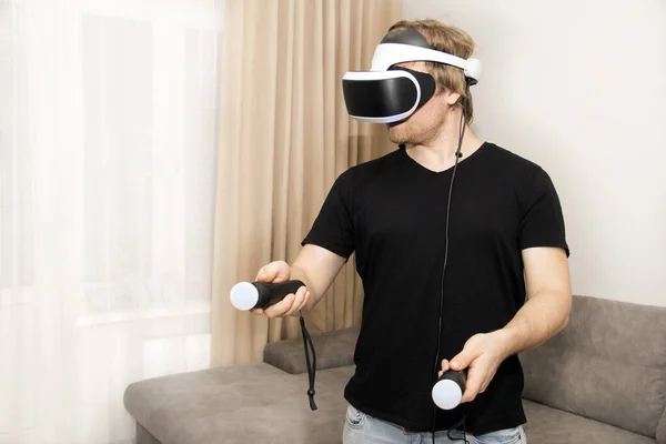 Um homem vestindo um fone de ouvido de realidade virtual e segurando controladores joga um jogo de vídeo em casa. Uma pessoa usando uma tecnologia moderna de fone de ouvido de realidade virtual. — Fotografia de Stock