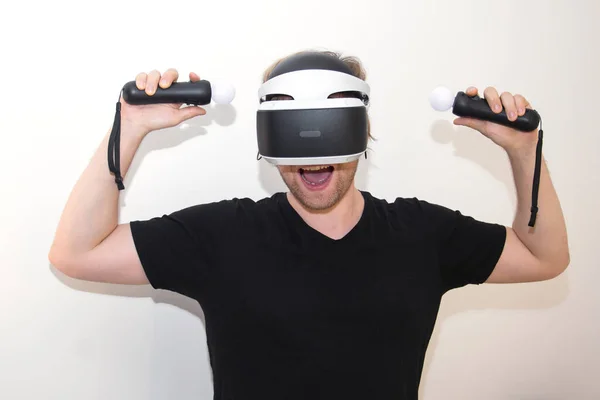 Een jongeman speelt VR. De joysticks. Witte achtergrond. Afbeelding van een vrolijke bebaarde jongeman in een virtual reality apparaat, staand op een witte achtergrond en met joysticks. Stockafbeelding