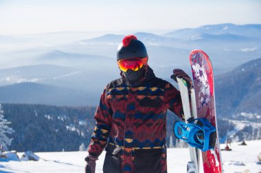 Dağın tepesinde snowboard kıyafeti giymiş bir adam. Dağların tepesinde elinde bir snowboard 'la duran genç adam.