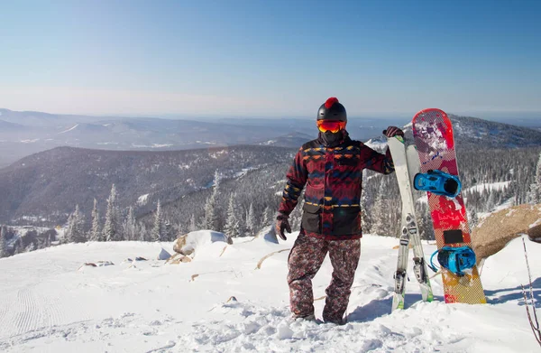 Egy férfi snowboardfelszerelésben egy hegy tetején. Fiatalember áll egy snowboarddal a hegyek tetején — Stock Fotó