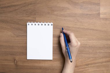 Ahşap bir masada boş bir sayfada, açık bir defterin yanında yazmaya hazır mavi bir kalem tutan genç beyaz bir el. Yukarıdan görüntüle.