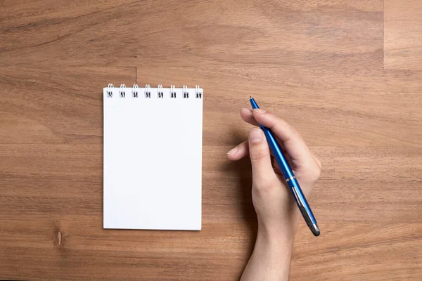 Ahşap bir masada boş bir sayfada, açık bir defterin yanında yazmaya hazır mavi bir kalem tutan genç beyaz bir el. Yukarıdan görüntüle.