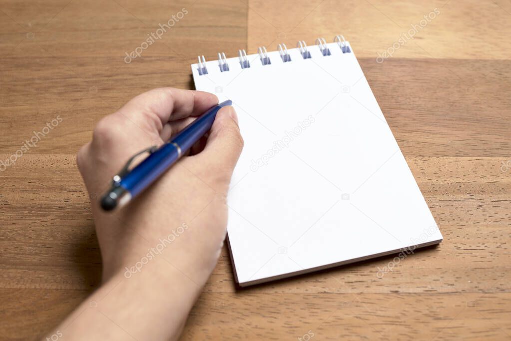 Left hand of a young left-handed caucasian woman writing with a blue pen on a blank page of an open notebook on a wooden desk. Subjective point of view.