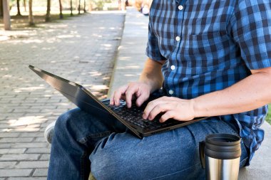 Dışarıda dizüstü bilgisayarda yazan tanınmamış genç adam. Yanında alüminyum bir termal bardak var. Dijital göçebe kavramı, açık havada çalışmak veya çalışmak.