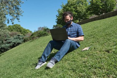Parkta oturan bir dizüstü bilgisayarla çalışan ve ders çalışan bez maskeli genç bir adam. Dijital göçebelik kavramları ve koronavirüsten korunma, Covid salgını sırasında.