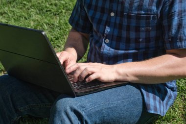 Güneşli bir günde çimlerin üzerinde oturmuş dizüstü bilgisayarda yazan tanınmamış genç adam. Dijital göçebe kavramı, dışarıda çalışma veya öğrenim.