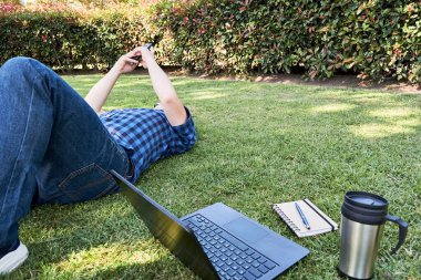Çimlerin üzerinde cep telefonu kullanarak uzanan, ders çalışmaktan ya da dışarıda çalışmaktan dinlenen tanınmamış genç bir adam. Yanında dizüstü bilgisayarı, kalemle birlikte bir defter ve yeniden kullanılabilir çevre dostu bir termal bardak var..