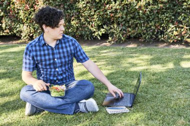 Genç adam çalışırken ya da bilgisayarında çalışırken çimenlerde oturmuş taze salata yiyor. Açık havada çalışma, sağlıklı yaşam ve yeme kavramları.