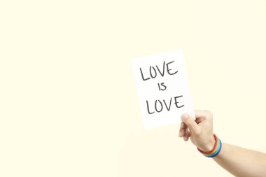 Gökkuşağı bileziği olan genç bir el, LGBT sembolü, elinde el yazısıyla yazılmış bir mesaj olan bir kağıt parçası: aşk aşktır. Aşk kavramları, eşitlik ve ayrımcılık karşıtlığı. Kopyalama alanı olan resim.
