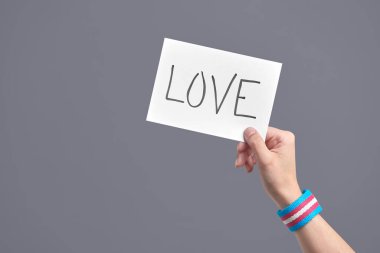 Young hand with a transgender flag bracelet holding a piece of paper with a handwritten message: love. Concepts of love, equality and non-discrimination. Image with copy space.