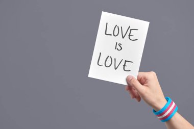 Young hand with a transgender flag bracelet holding a piece of paper with a handwritten message: love is love. Concepts of love, equality and non-discrimination. Image with copy space.