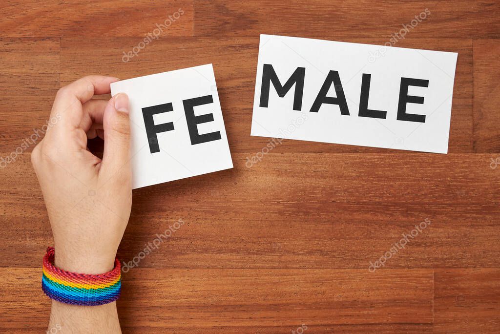 Hand changing the word male into female or the opposite, wearing a rainbow bracelet, colors of the lgbt pride flag. Conceptual image about gender identity and transgender. Copy space, wood background.