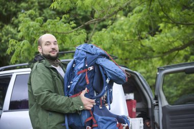 Adam sırt çantasını arabanın bagajından çıkarırken kameraya bakıyor. Kavram: açık hava tatillerinin keyfi..