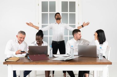 Bin yıllık başarılı Hint erkek patron CEO 'sunun grup fotoğrafı ofiste çok ırklı iş arkadaşlarıyla el kaldırarak arkasında duruyor. Masanın başında oturan çeşitli şirket iş adamları birlik ve liderlik kavramını gösteriyor.