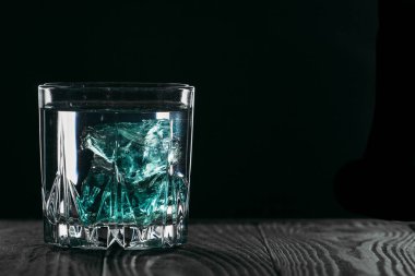 Siyah arka planda, tahta yüzeyinde mavi buz küpleri olan bir bardakta cin. Buzlu alkol. Bir yazıt için yer.