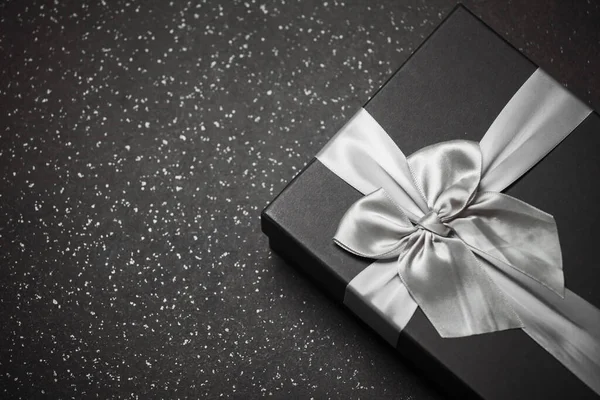 Black gift box with a silver ribbon and a large bow on a granite dark background. Place for the inscription