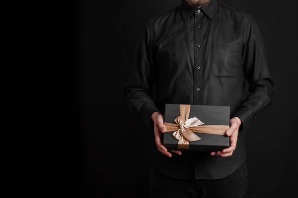 A guy in a black shirt is holding a gift with a golden ribbon. Black background. Black gift box in hands.