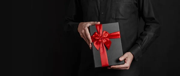 黒いシャツの男が赤いリボンの贈り物を持っている 黒の背景 ブラックギフトボックスを手に パノラマ写真 碑文の場所です バナー — ストック写真