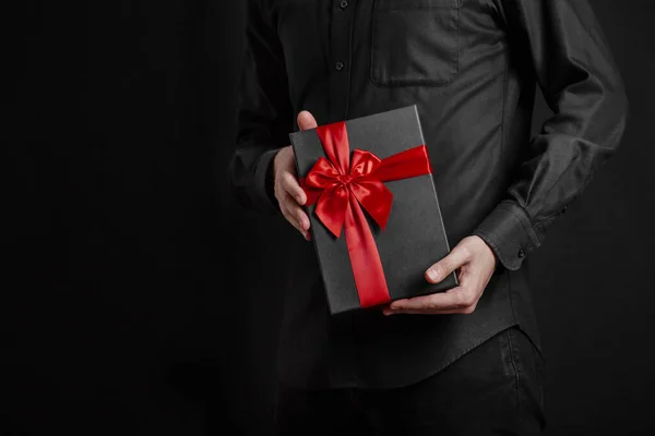 A guy in a black shirt is holding a gift with a red ribbon. Black background. Black gift box in hands.