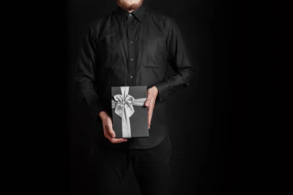 黒いシャツの男が赤いリボンの贈り物を持っている 黒の背景 手にブラックギフトボックス — ストック写真