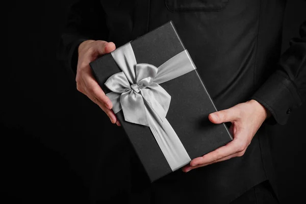 A guy in a black shirt is holding a gift with a red ribbon. Black background. Black gift box in hands. Close-up photo.