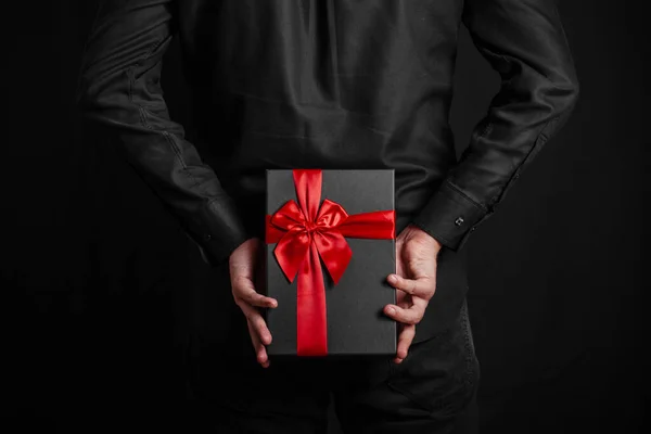 Man in a black shirt holds a gift with a red ribbon behind his back. Black gift box in hands. Surprise.