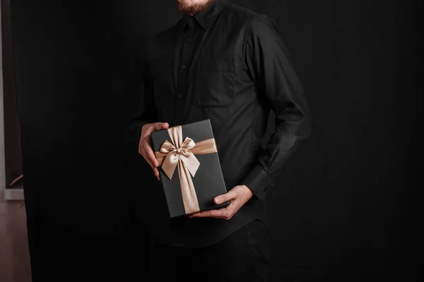黒いシャツの男がゴルドボリボンで贈り物を持ってる 黒の背景 手にブラックギフトボックス — ストック写真