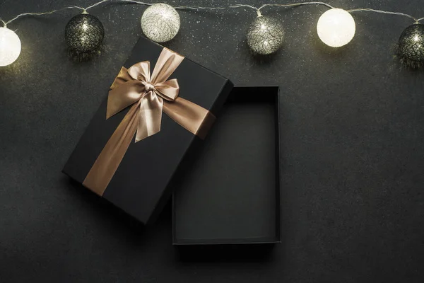 Opened black gift box with golden ribbon. Gift on a black granite surface. Christmas lights above the box.