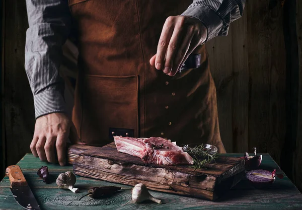 革のエプロンの男は骨とステーキに黒コショウを振りかける 木の板の上の生肉 — ストック写真