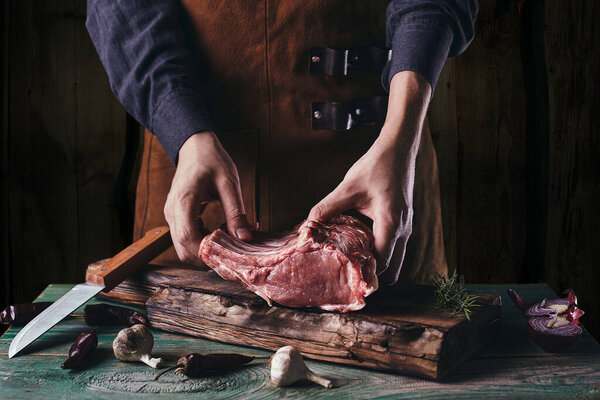 A guy in a leather apron is slicing raw meat. The butcher cuts the pork ribs. Meat with bone on a wooden cutting board.