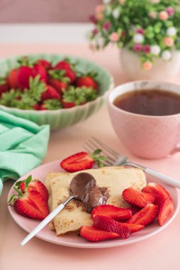 Çikolatalı krep ve taze çilek, pembe arka planda çay ile servis edilir.